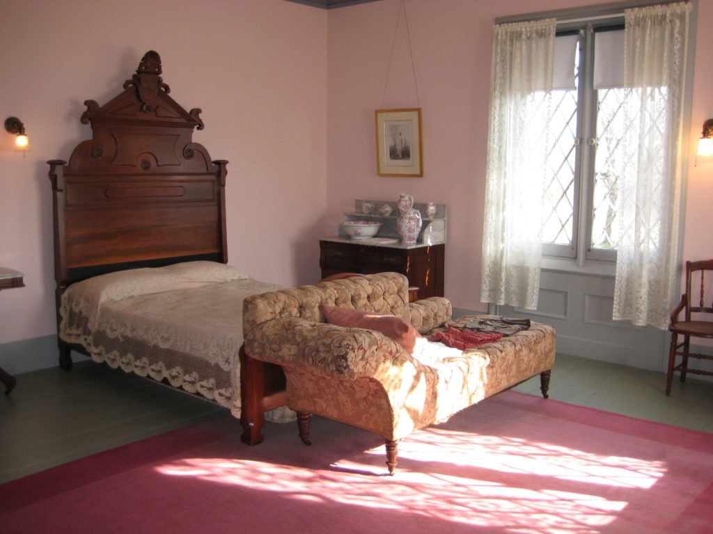 sun filled pink room with pink rug, bed with tall carved headboard, day bed at foot of bed