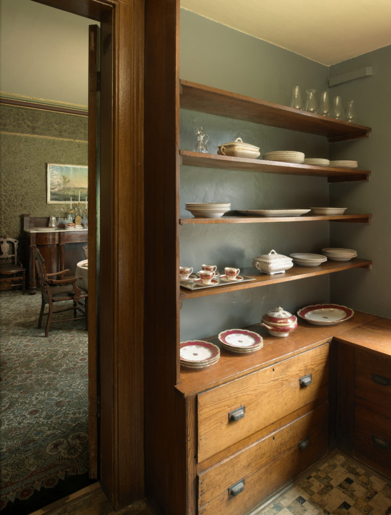 Small room with shelf holding dishes, open door to room with patterned carpet and elaborate wall covering.