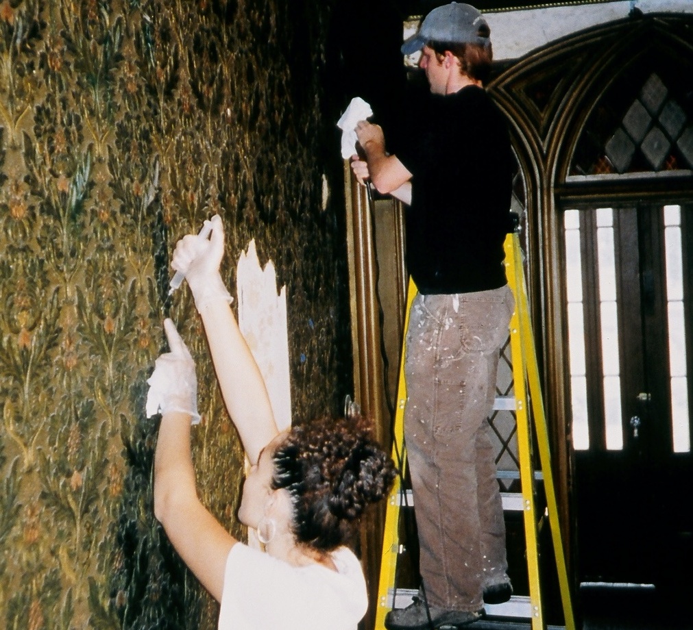 Two workers repair damaged wallcovering in entry hall, one injecting substance in damaged area to readhere lincrusta to the wall.