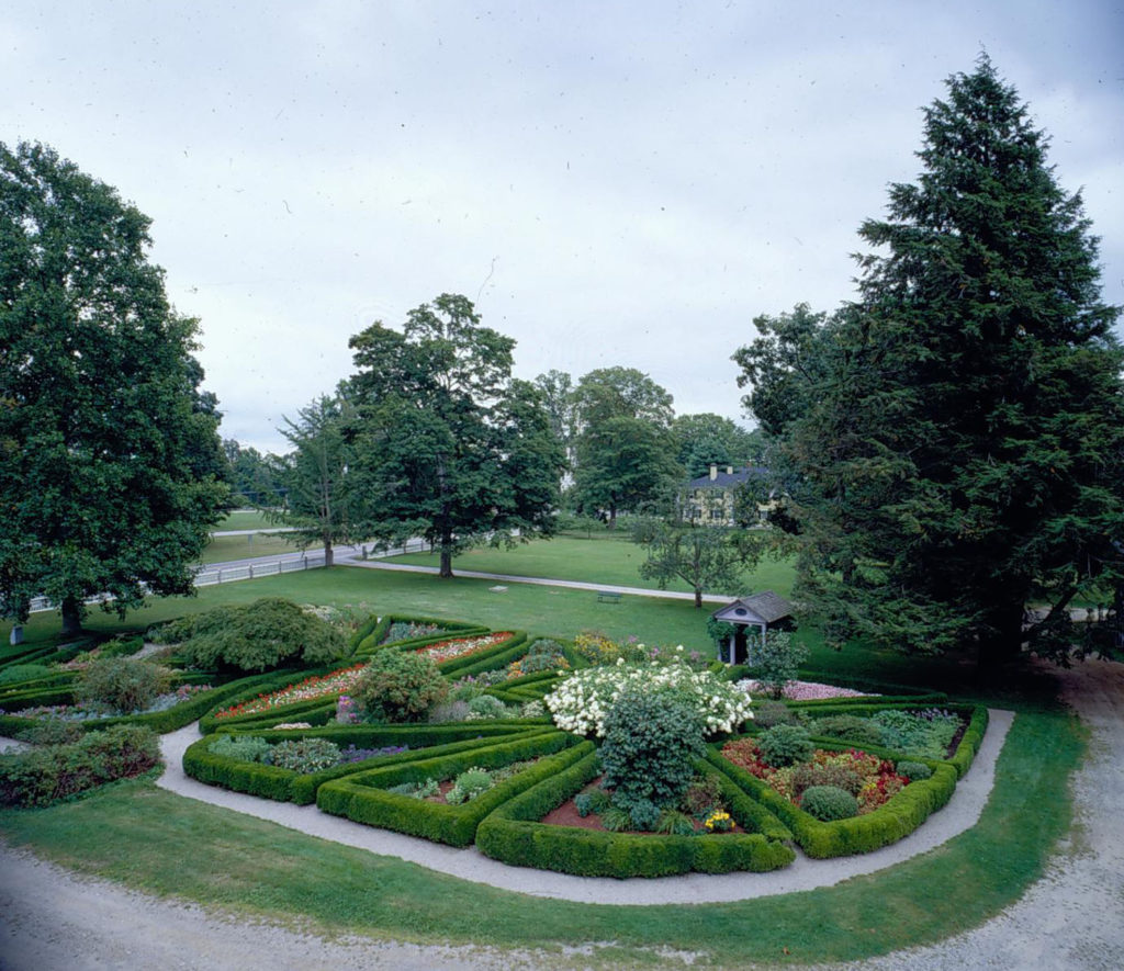Front lawn and garden seen from above, 21 colorfully planted flowerbeds grouped together, each edged with boxwood, with gravel paths between beds. 
