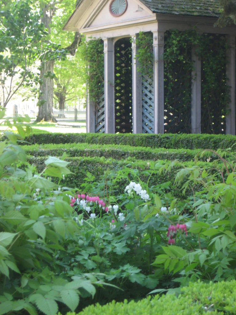 Wooden structure 9 by 12 feet, lattice sides, peaked roof, and parterrebed with bleeding heart flowers in foreground.