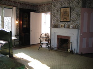 Blue patterned wallpapered room with green bed and chair, marble fireplace, and open door in corner in front of tall bookcase.
